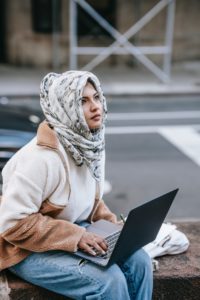 Woman with a hijab, sitting outside, with a laptop.